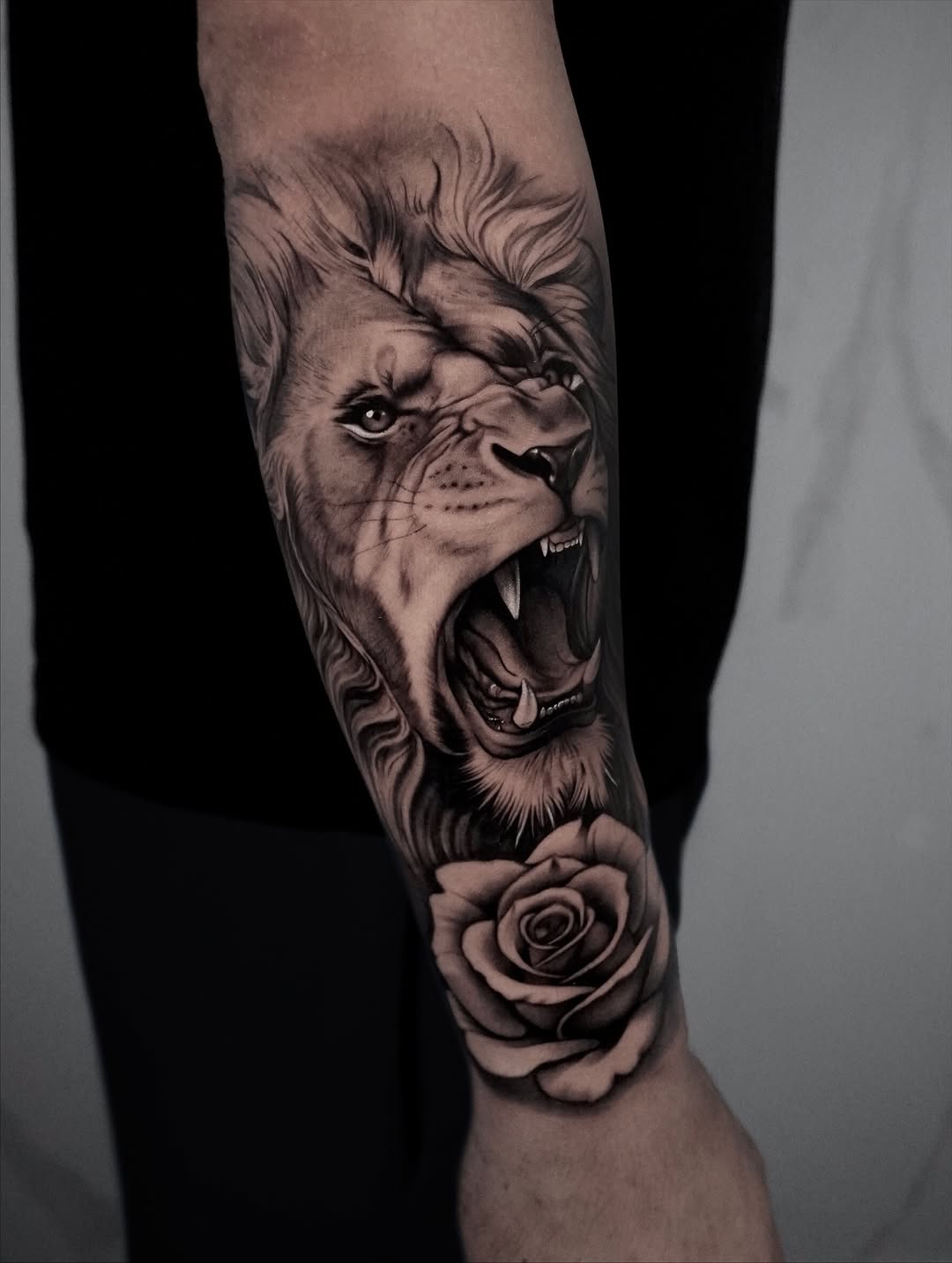 Roaring lion head with open mouth and rose flower tattoo on forearm in grayscale ink.