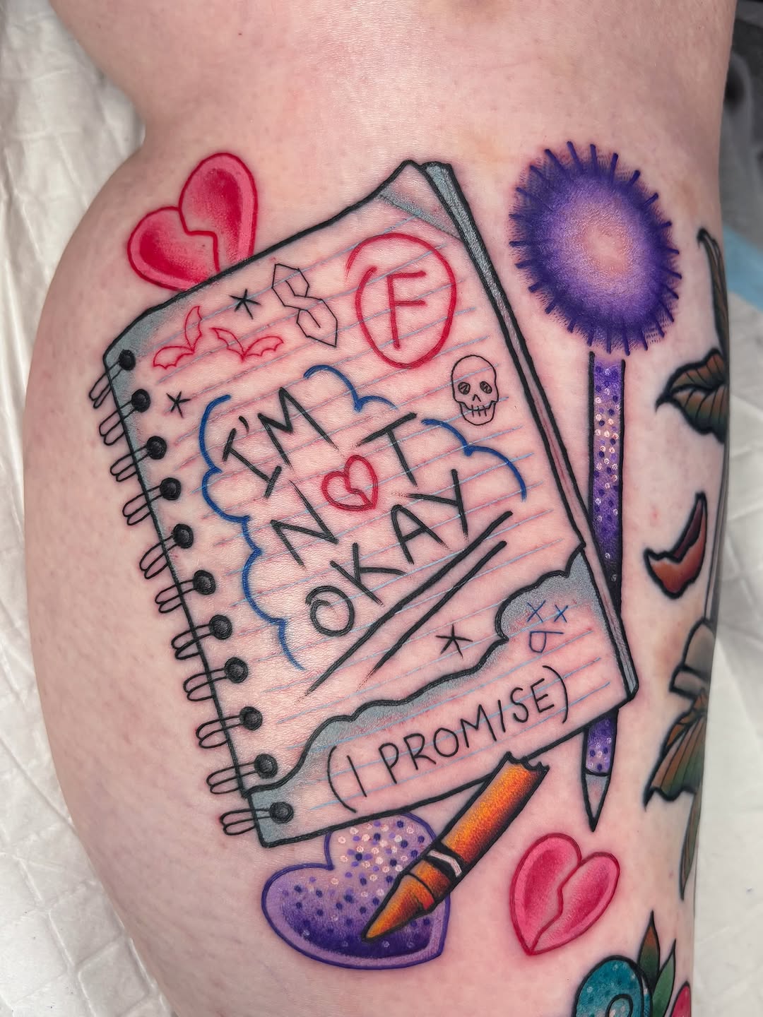 Colorful notebook tattoo on leg featuring spiral-bound pages with handwritten text, hearts, skull, brush, and decorative elements in red, blue, and purple ink.