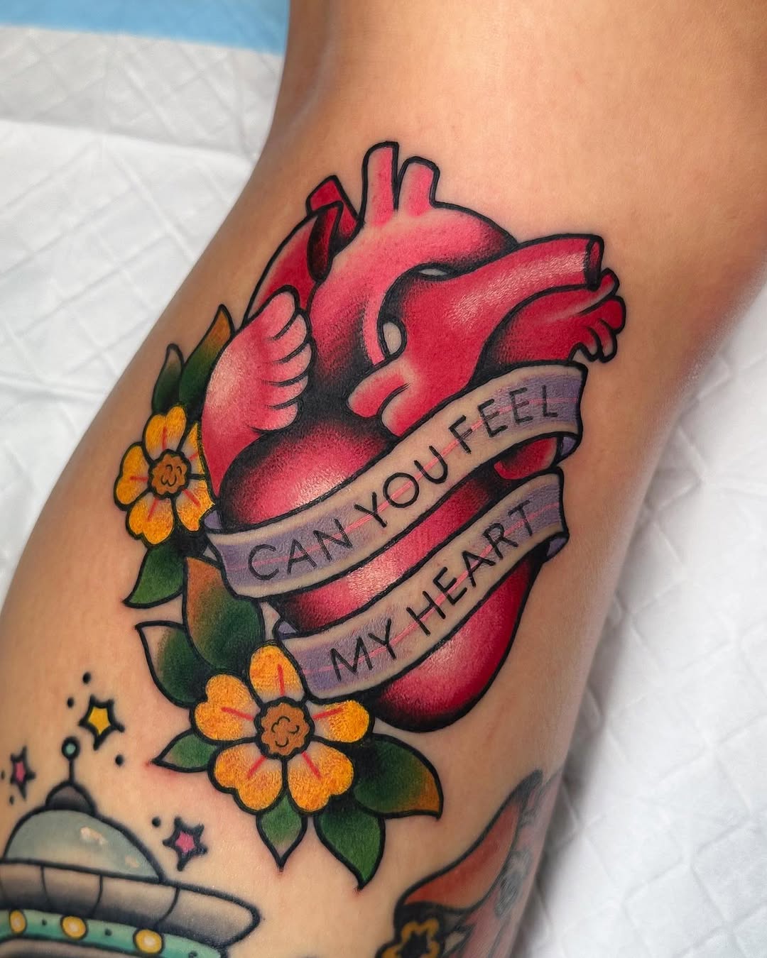 Anatomical heart in red with yellow flowers and a banner reading "Can you feel my heart" tattooed on an arm.