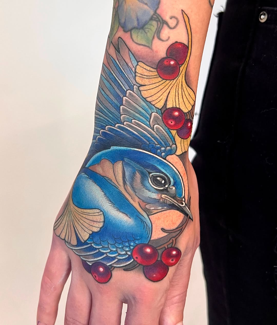 Colorful hand tattoo featuring a blue bird with spread wings surrounded by red berries and flowering branches in yellow and green tones.