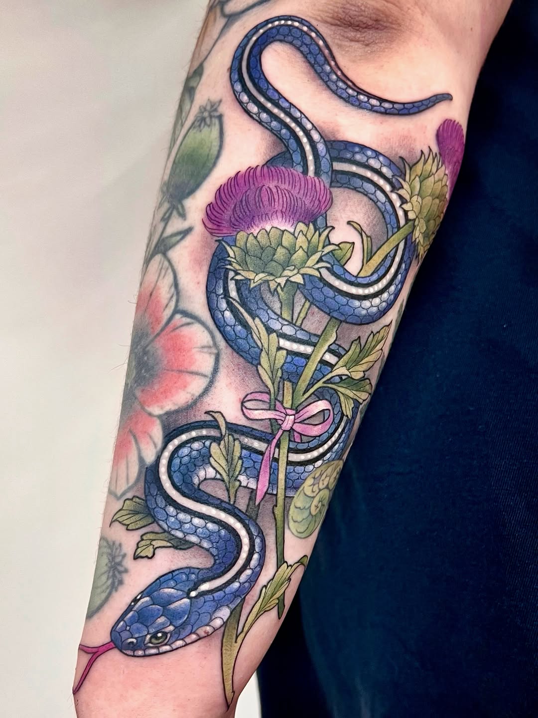 Blue snake intertwined with purple thistle flowers and green leaves with a pink bow, tattooed on the forearm.