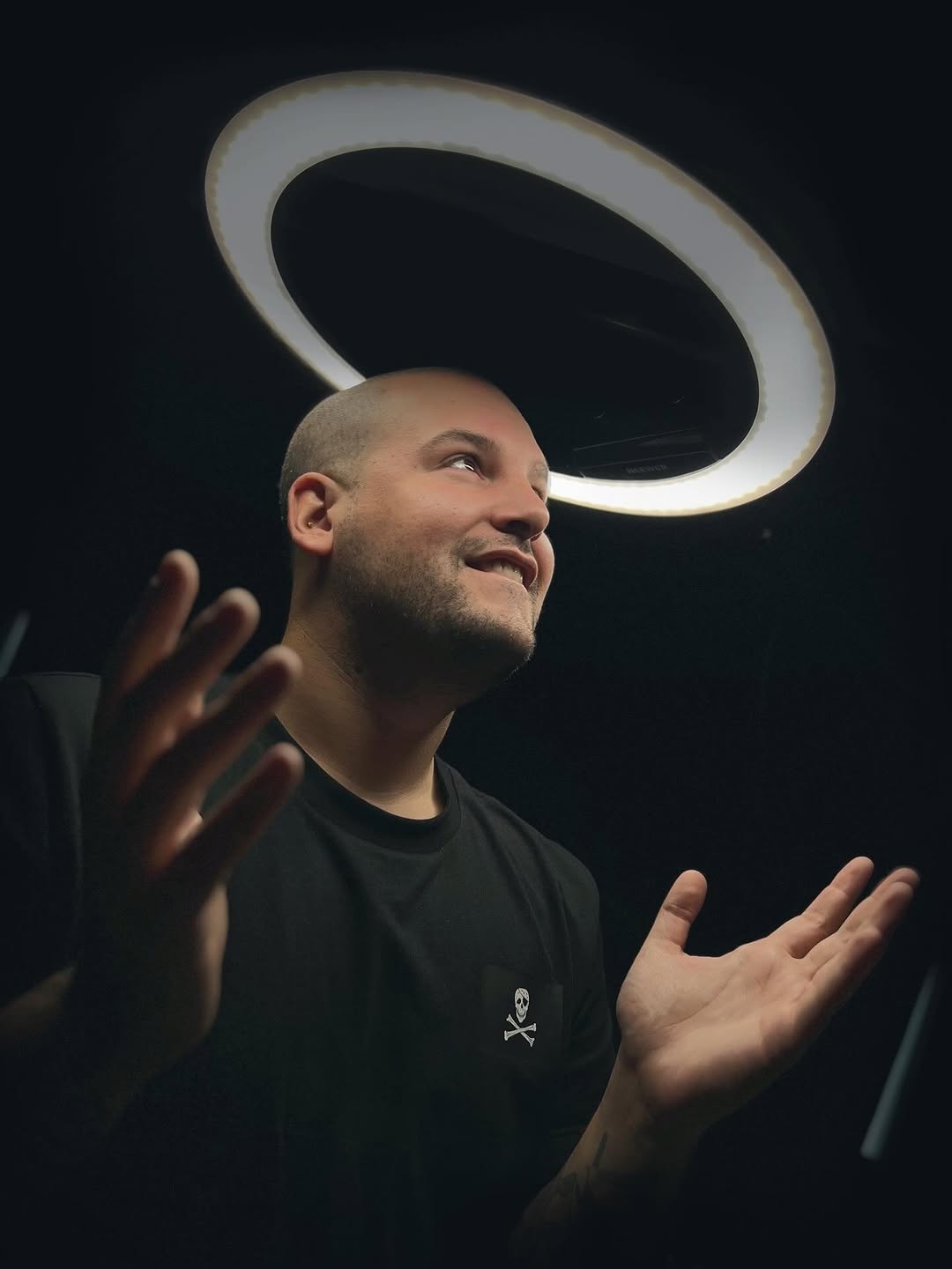 Man wearing black t-shirt with small skull and crossbones chest tattoo, photographed against dark background with circular light fixture overhead.