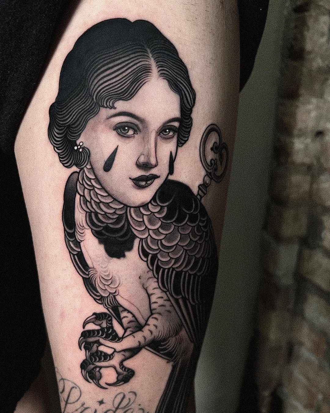 A black and white shoulder tattoo featuring a woman with wavy hair, decorative collar, and bird wings, holding taloned feet.