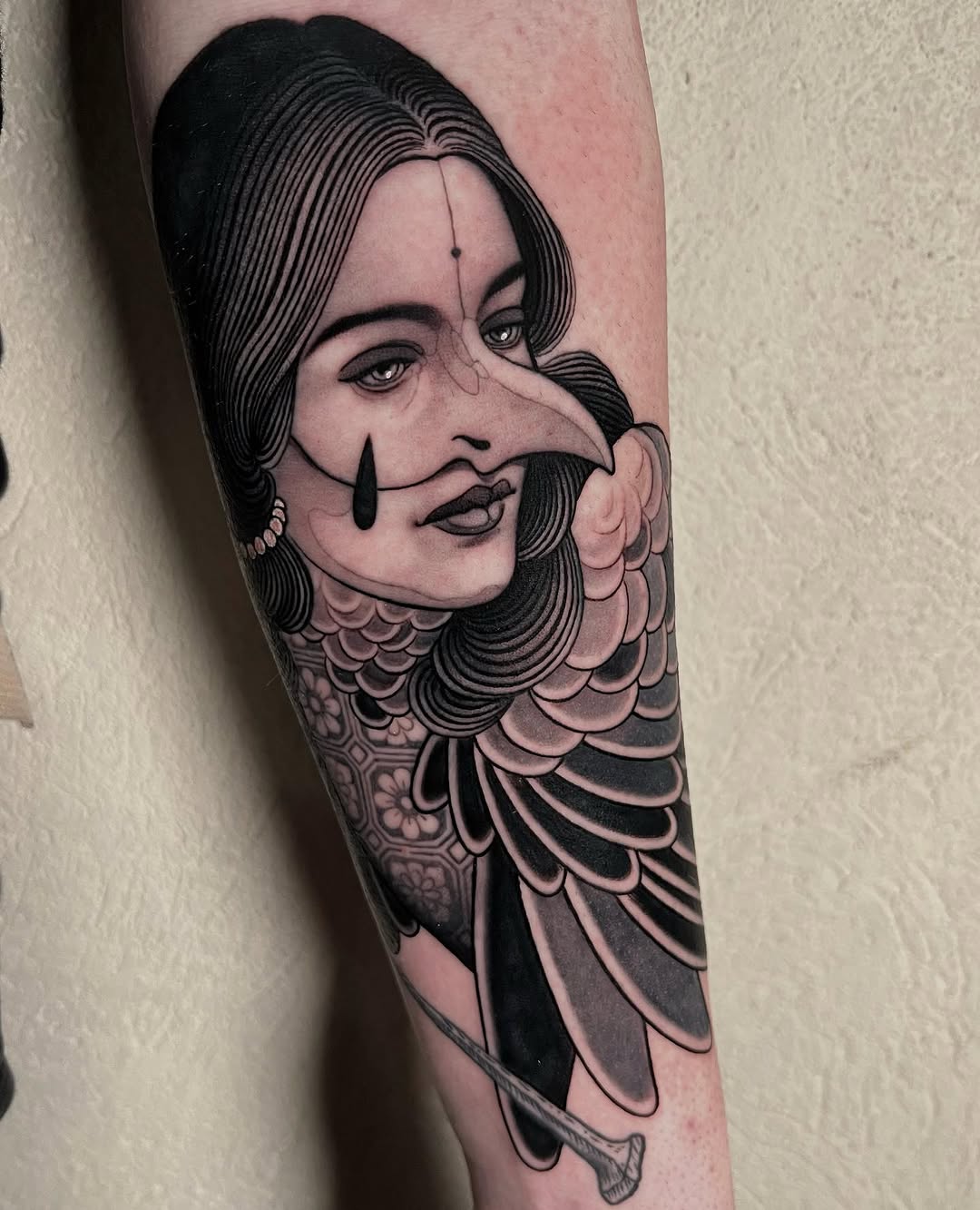 Surreal female portrait tattoo with elongated nose, teardrop marks, and feathered wing shoulders in black and gray ink on forearm.