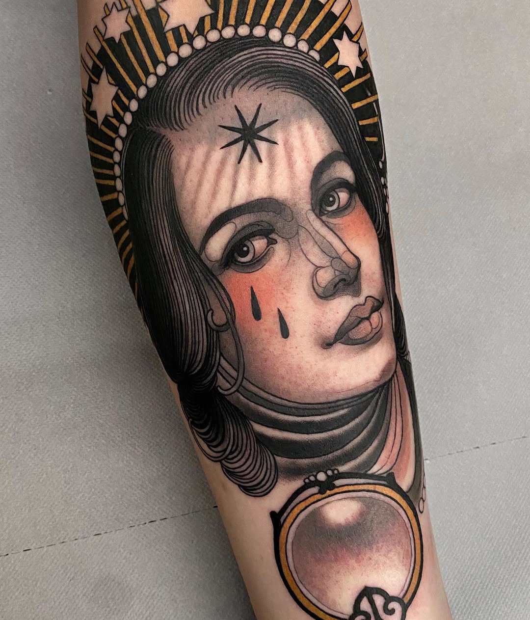 Portrait tattoo of a woman's face with a star on her forehead, pearl halo, tear marks, and decorative orb below, rendered in black ink with gold and red accents on the forearm.