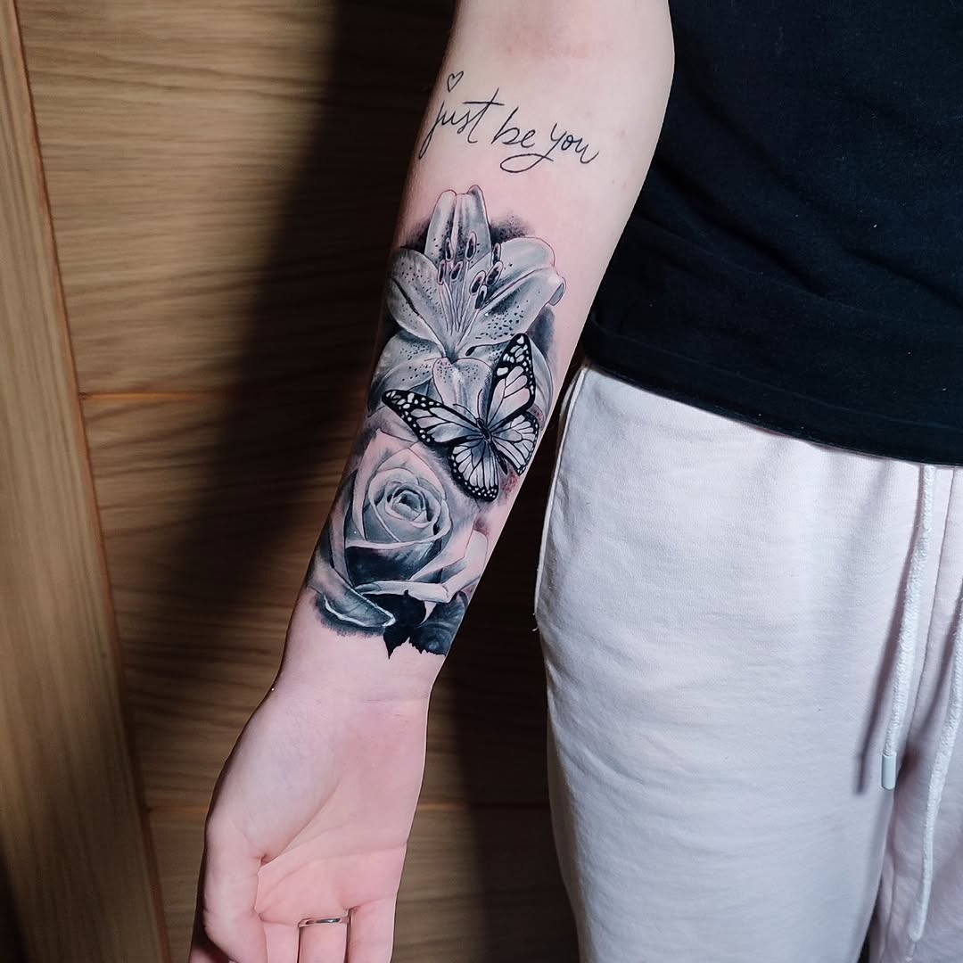 Forearm tattoo featuring a rose, lily, and butterfly with cursive text reading "just be you" and a heart symbol above the flowers.