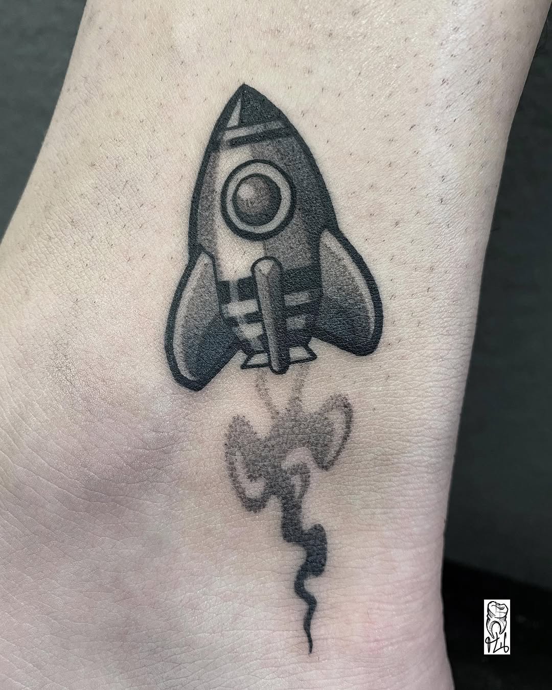 Rocket ship tattoo with circular window and flame exhaust on inner arm, rendered in black ink with shading and line work.