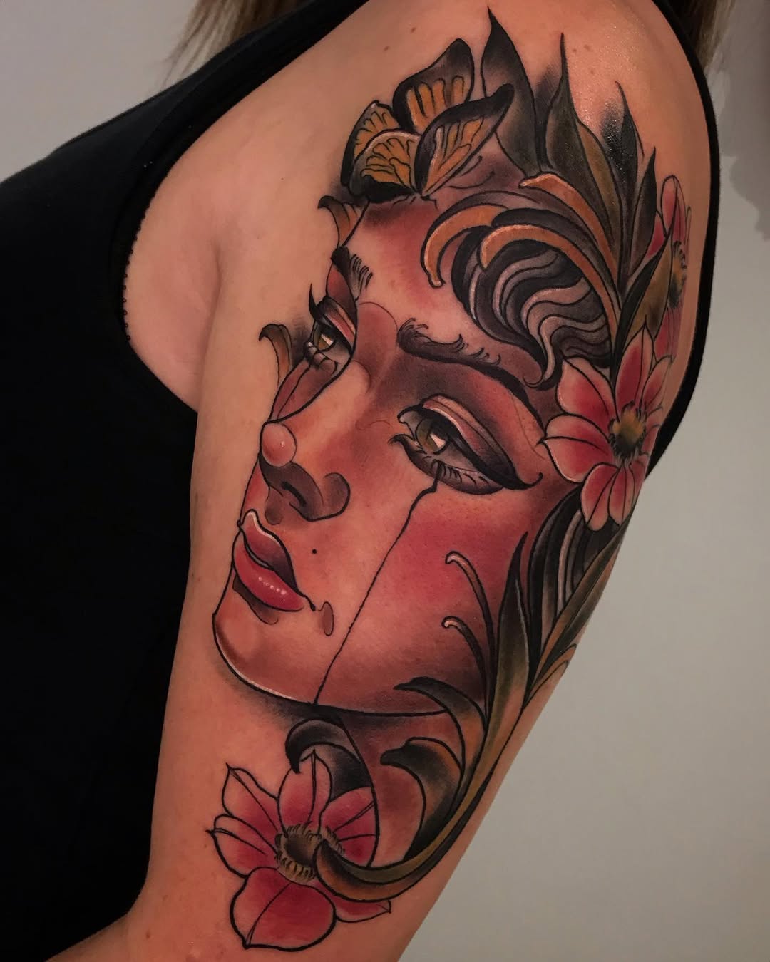 A woman's upper arm tattoo featuring a red-faced female profile surrounded by ornamental foliage, butterflies, and flowers in red and black ink.