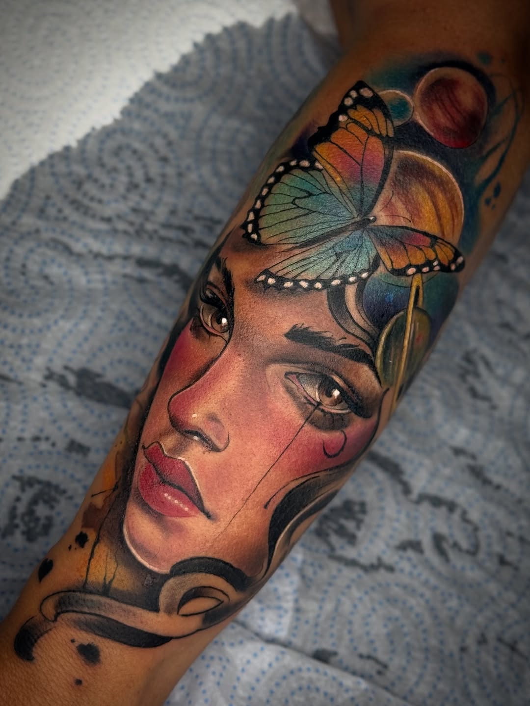 Colorful portrait tattoo on forearm featuring a woman's face with a butterfly on her head, rendered in orange, green, blue, and red tones.