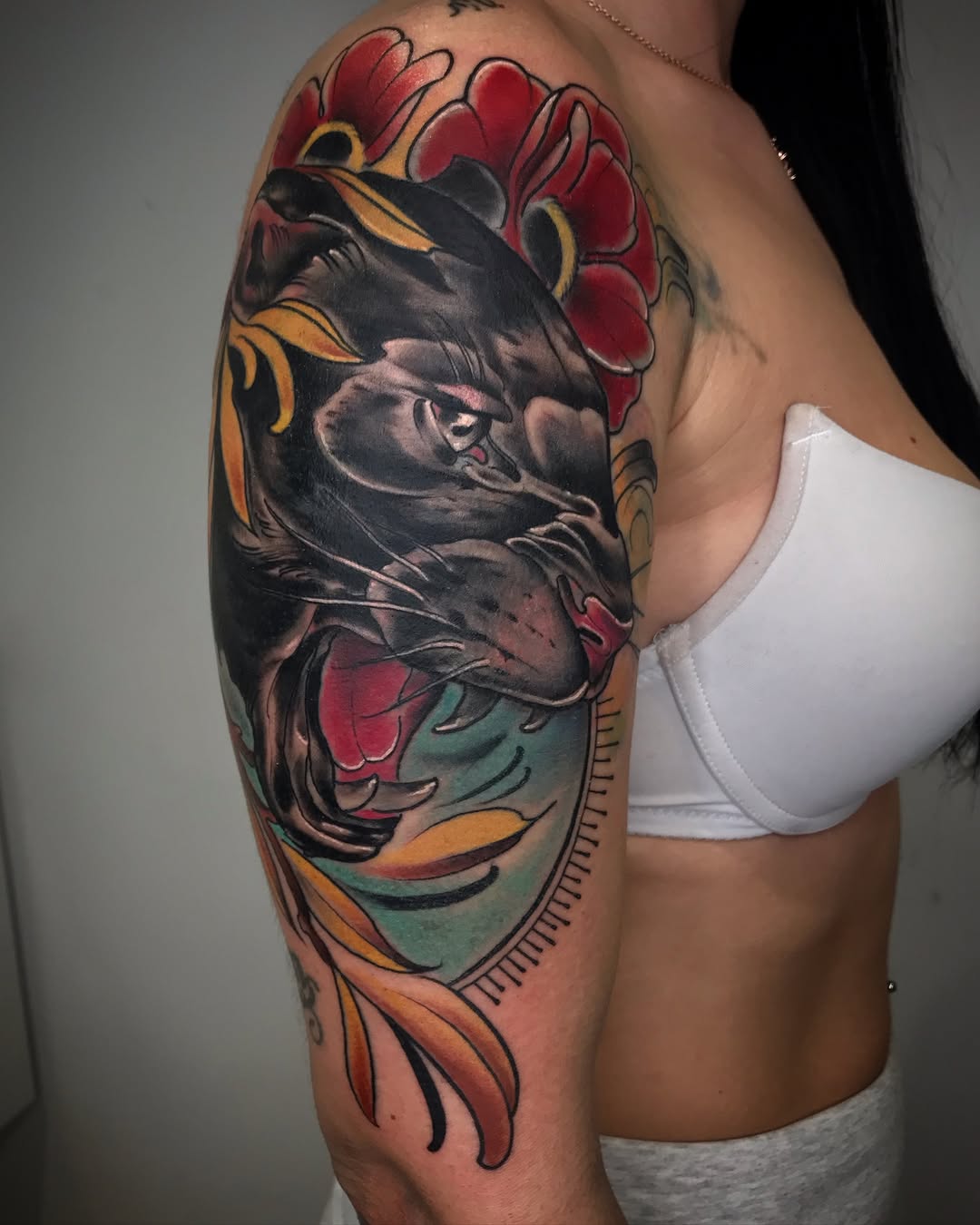 Colorful shoulder and upper arm tattoo featuring a detailed face surrounded by red flowers, green and orange decorative elements, and ornamental lines.