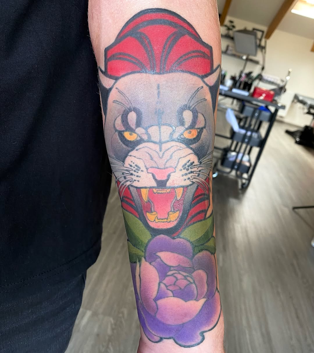 Colorful lion head tattoo on forearm featuring red mane, yellow eyes, green collar, and purple flower elements below.