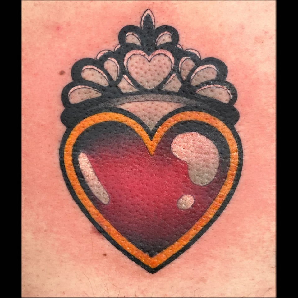Crowned heart tattoo with black outline, orange border, and deep red fill on skin with highlights and shading.