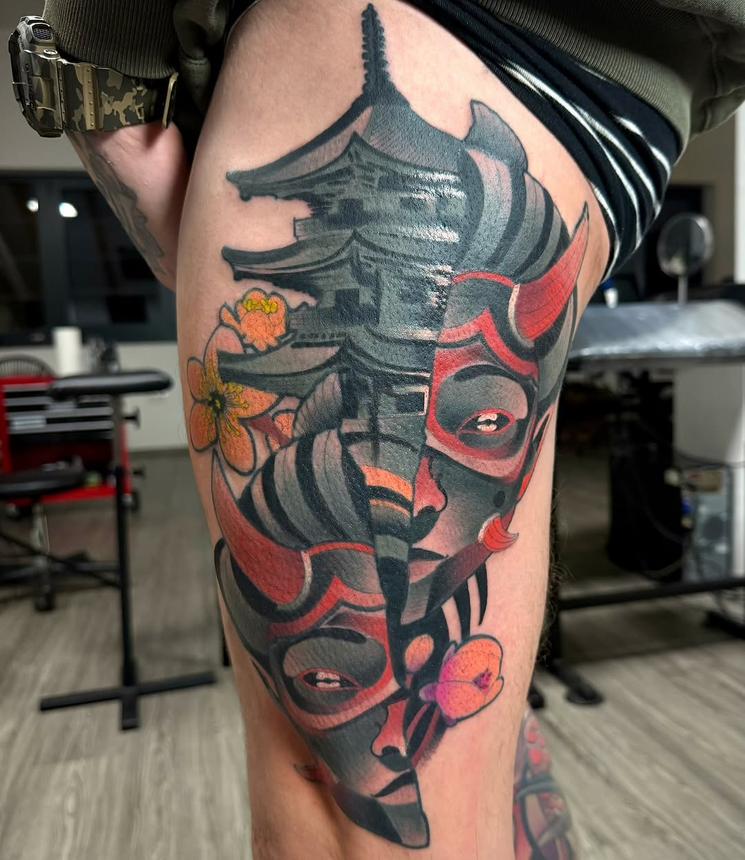 Colorful thigh tattoo featuring stacked demon and oni masks with flowers, rendered in red, orange, and dark grey on pale skin.