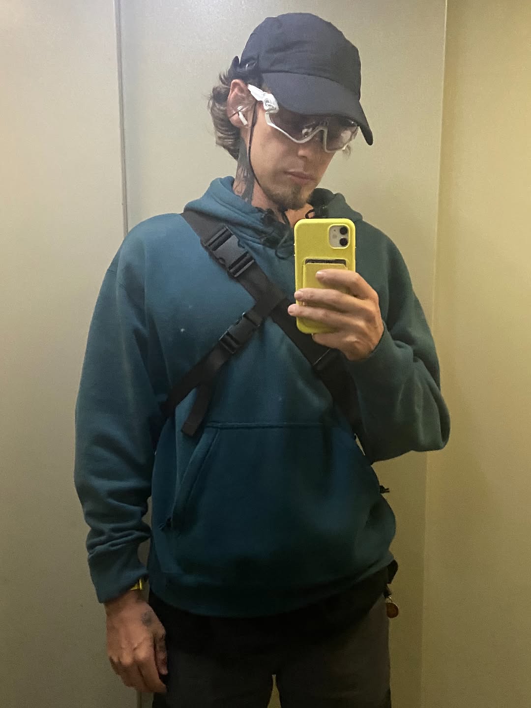 Person wearing a teal hoodie and black cap holds a yellow phone case, displaying a visible tattoo on their neck in black ink.