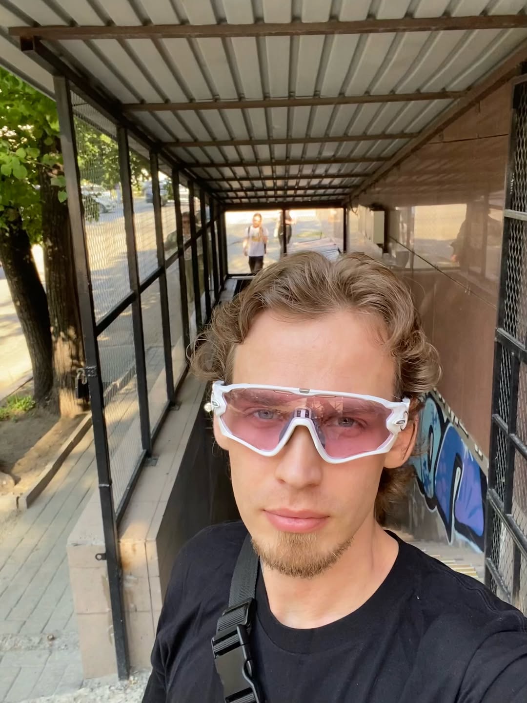 A person wearing white and pink sunglasses and a black shirt stands in a covered transit shelter with metal mesh panels.