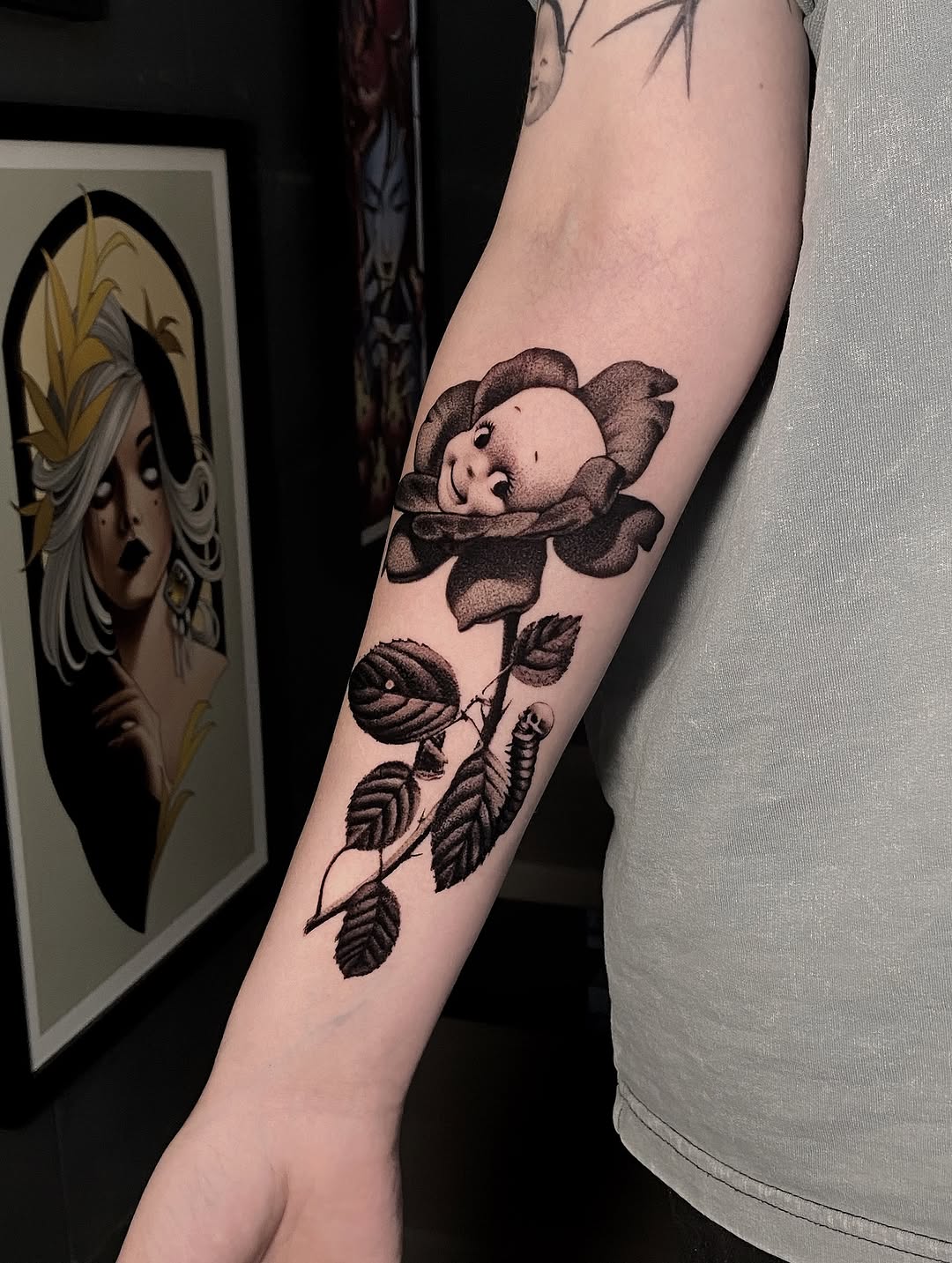 Baby face flower tattoo on forearm with leafy stem in brown ink, displayed on pale skin next to framed artwork.