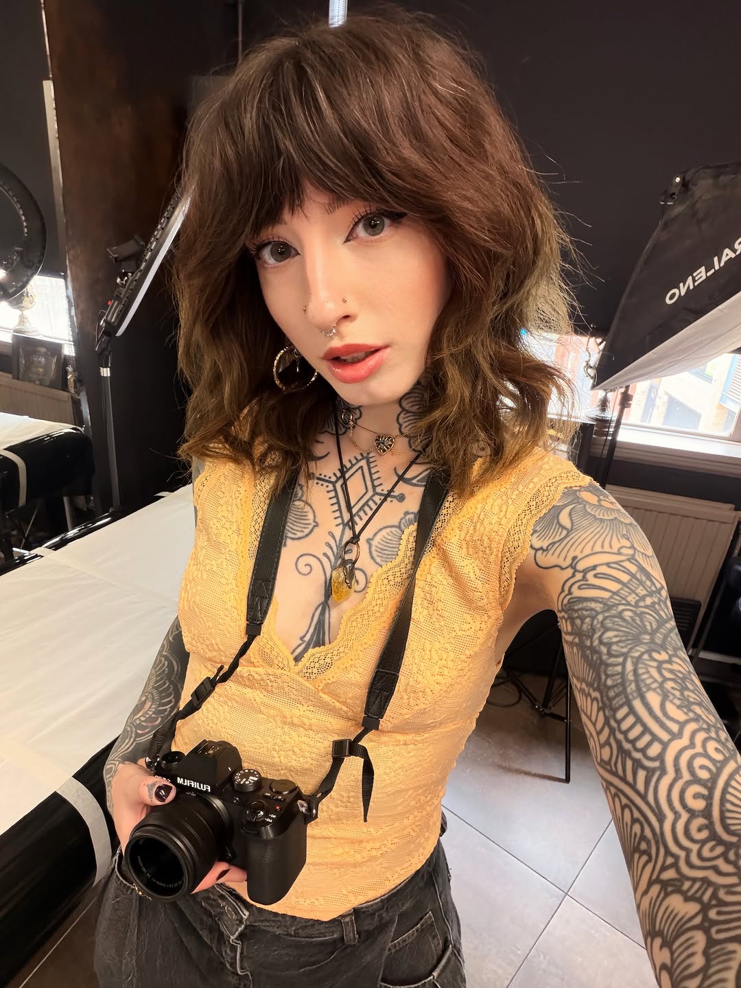 Person with brown wavy hair holding a camera, displaying detailed geometric and ornamental tattoos covering both arms and chest in black ink.