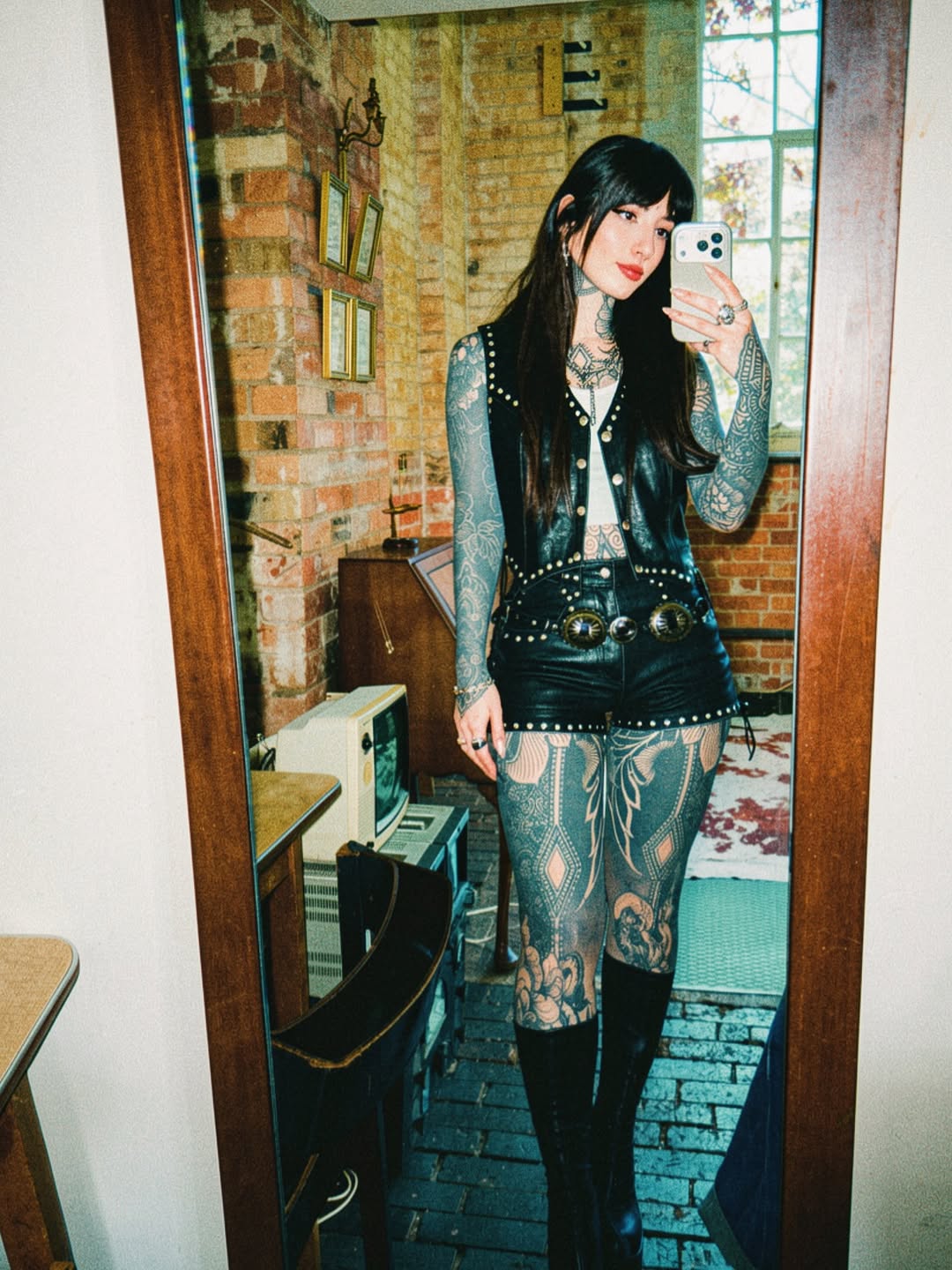 Woman with extensive arm and leg tattoos in black ink featuring floral and ornamental designs, wearing studded black leather vest and patterned stockings.