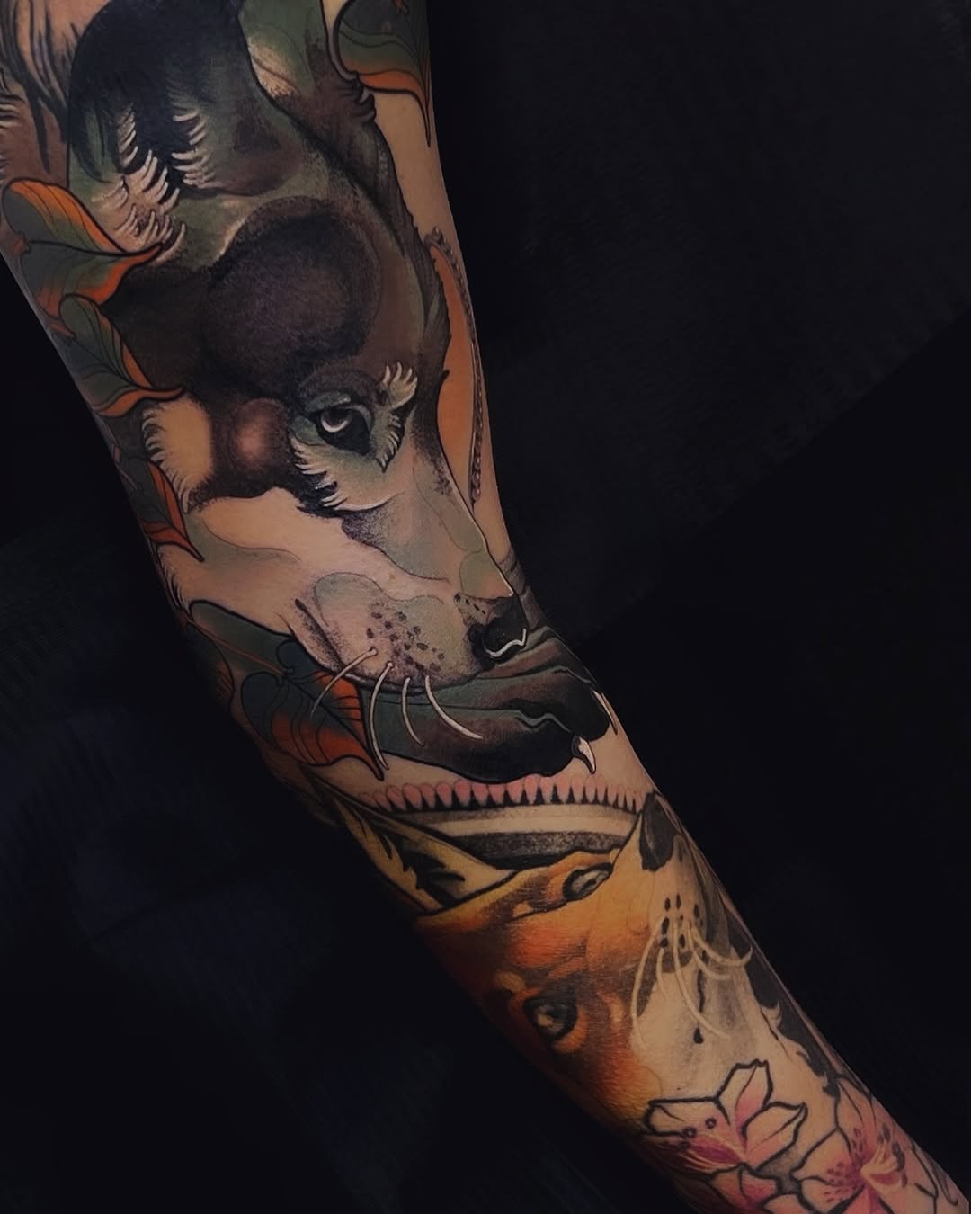 Colorful arm tattoo featuring stacked animal faces including a deer, fox, and fish with botanical elements in orange, black, and natural tones.