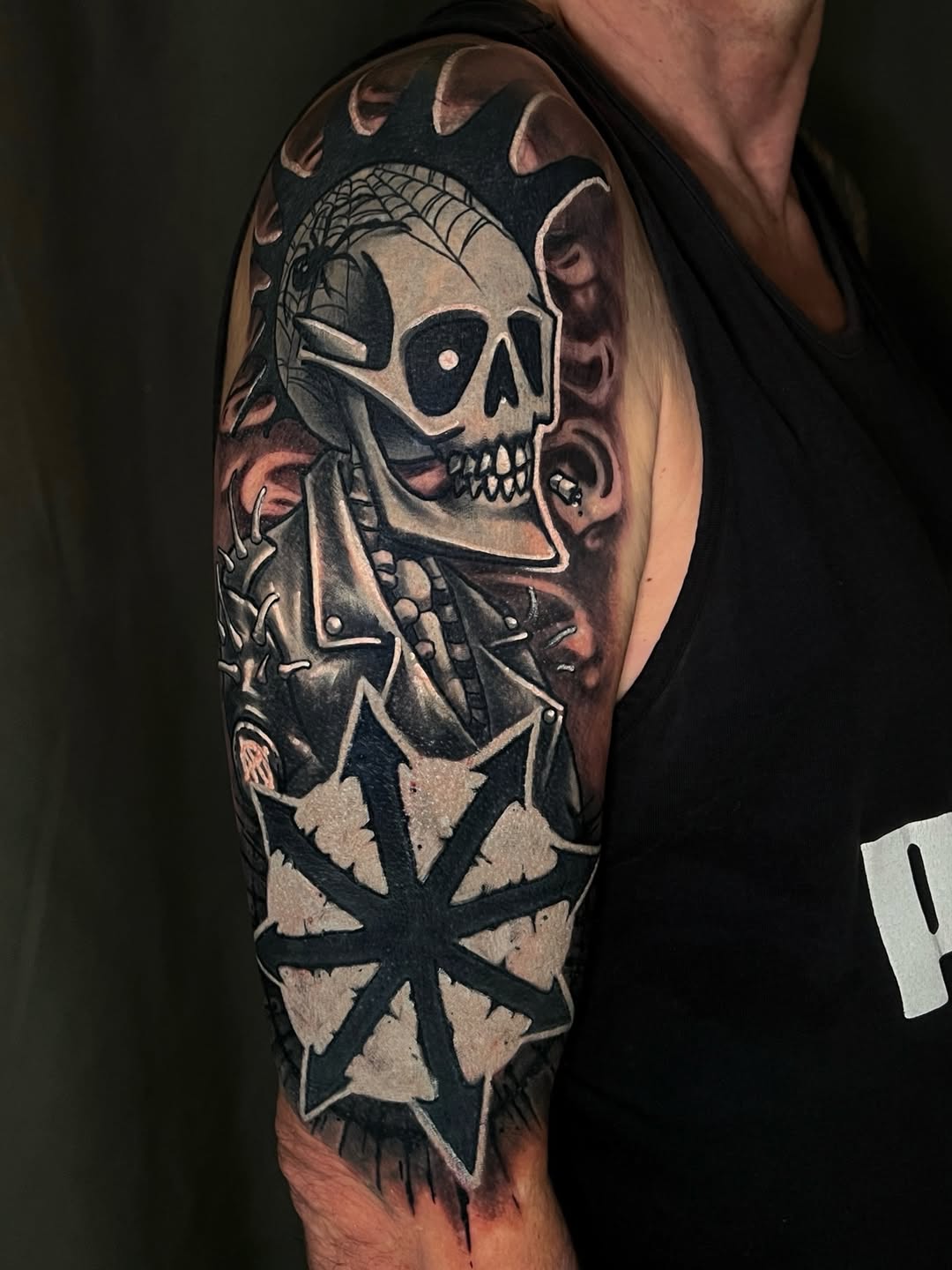 Skull with skeletal hand and geometric cross design tattooed on upper arm in black and grey tones with shading.