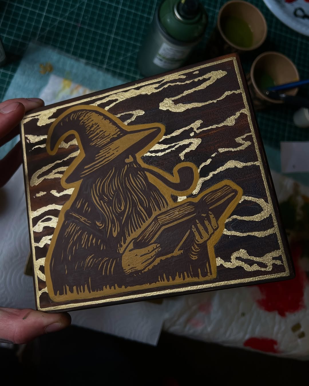 A wizard holding a wand wearing a pointed hat against a cloudy sky background, rendered in brown and gold tones on a wooden box.