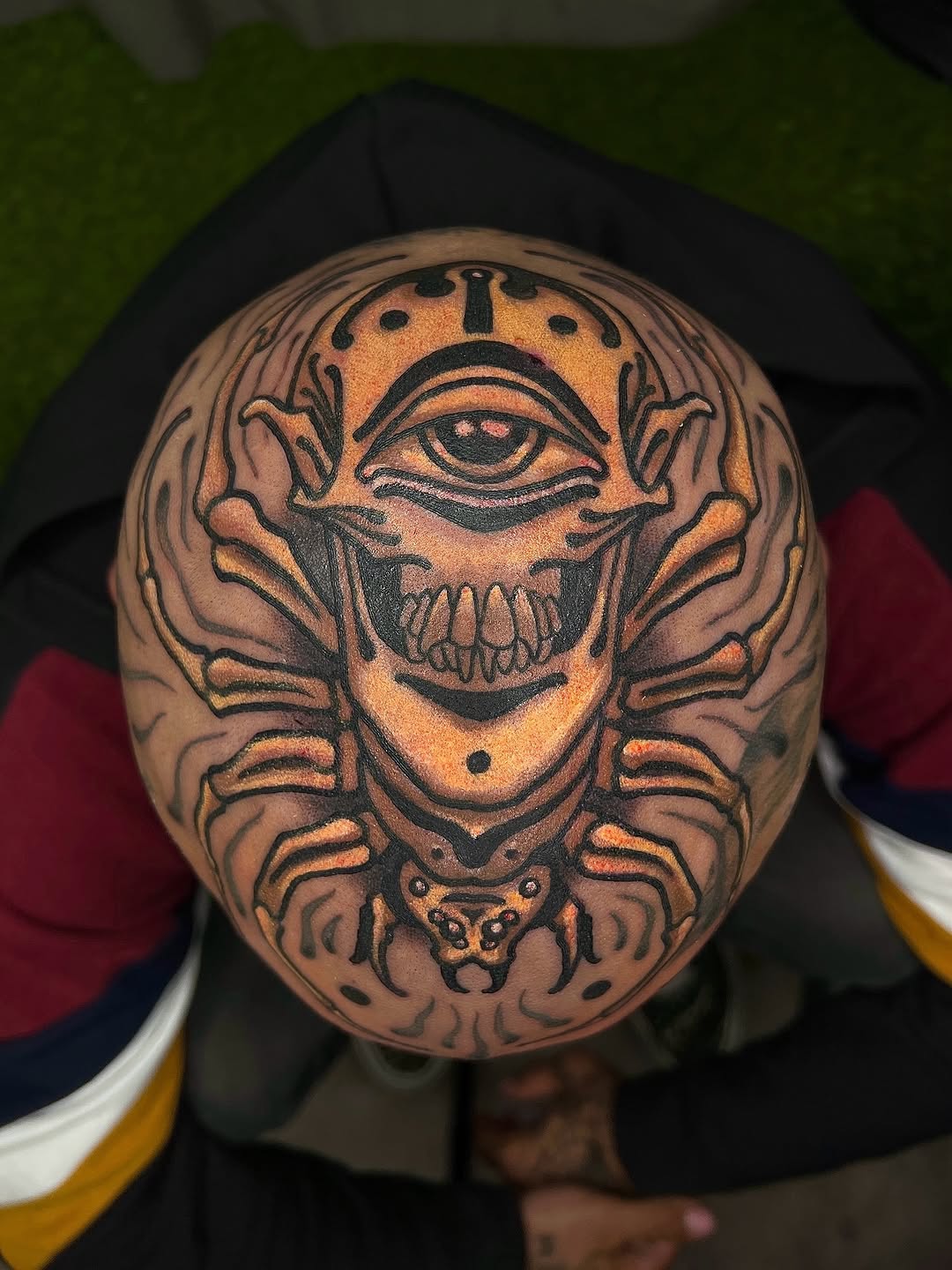 Skull with all-seeing eye and crab claws in orange and black ink on shaved head, displayed in portfolio setting.