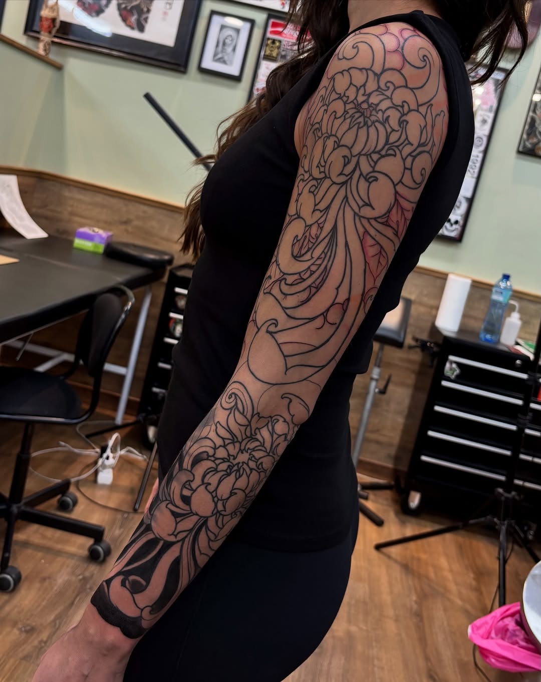 Full-sleeve arm tattoo featuring ornate scrollwork and floral designs in black ink with reddish undertones, displayed in a tattoo studio setting.