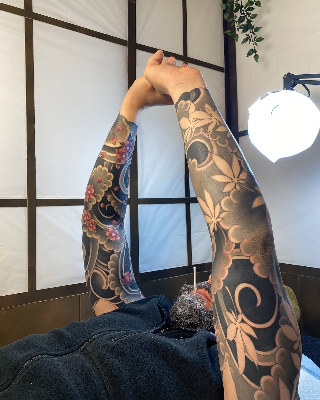 Detailed arm sleeve tattoos featuring ornamental flowers and botanical elements in black, gold, and red on both forearms displayed against a gridded window.