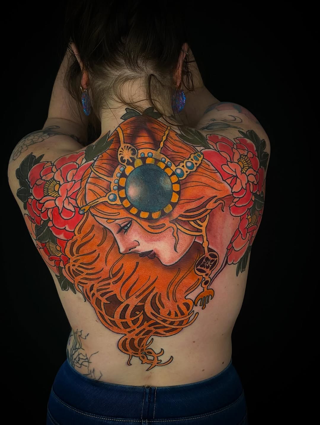 Back tattoo featuring a bearded man's profile with orange hair and beard, surrounded by red flowers and a large blue circular jewel ornament.