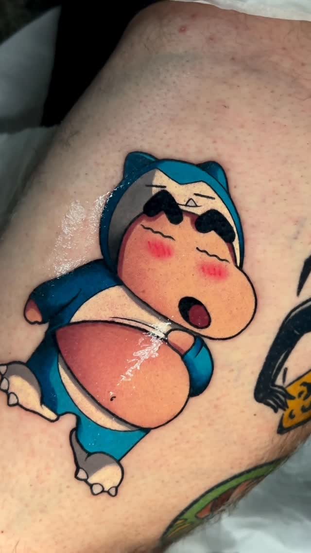 Colorful tattoo of a blue and peach-colored character with closed eyes and rosy cheeks, positioned on the upper arm.