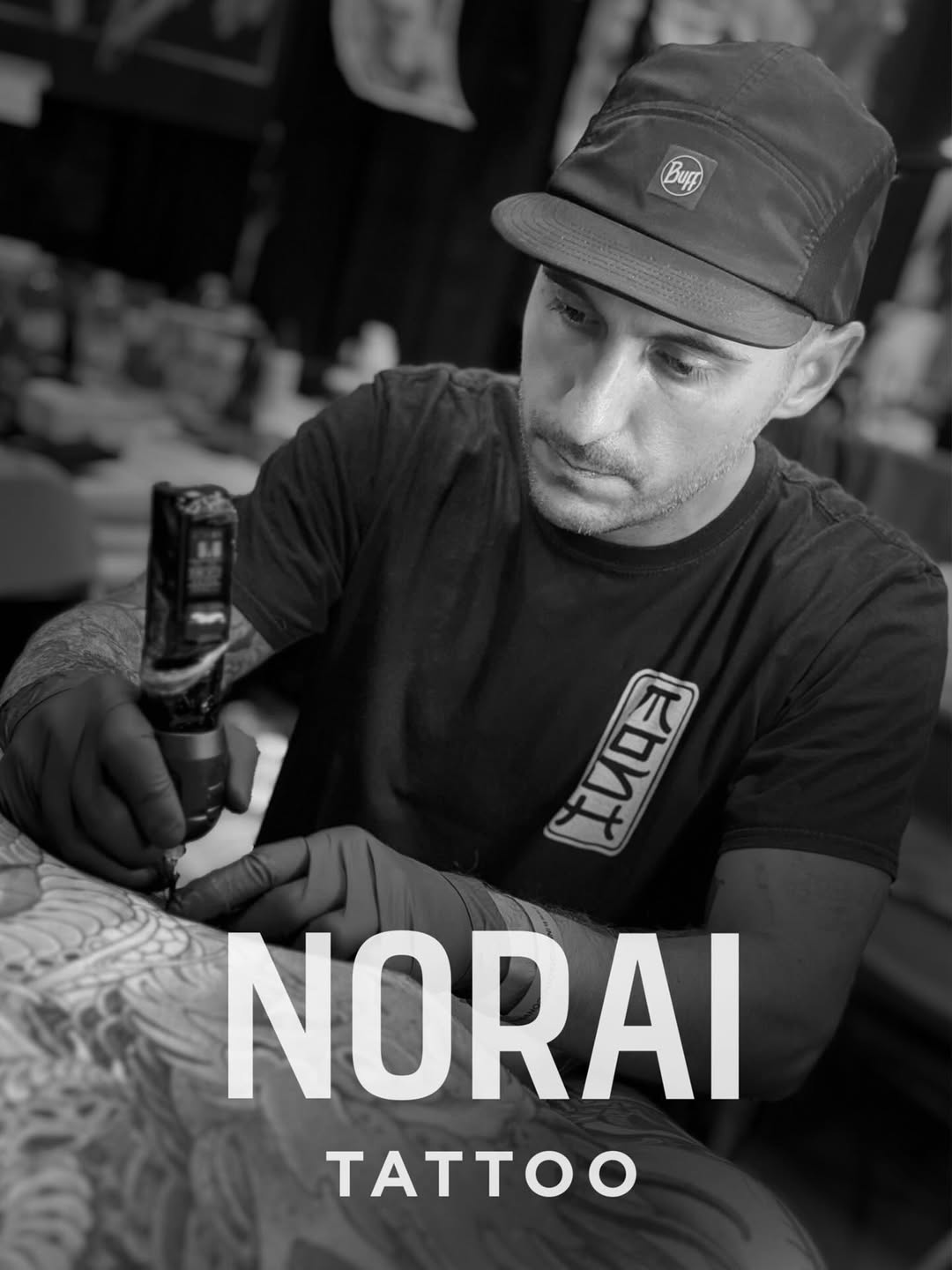 Tattoo artist in black Buff cap and shirt applies ink to client's leg during studio session, focusing intently on detailed work.