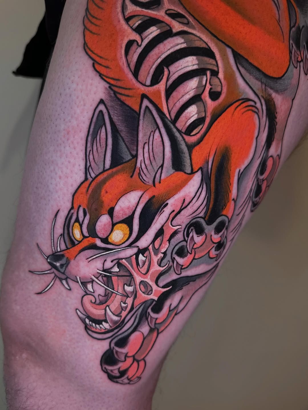 Colorful cat skull tattoo on thigh featuring orange and pink tones with yellow eyes, ornate decorative elements, and black outlines.
