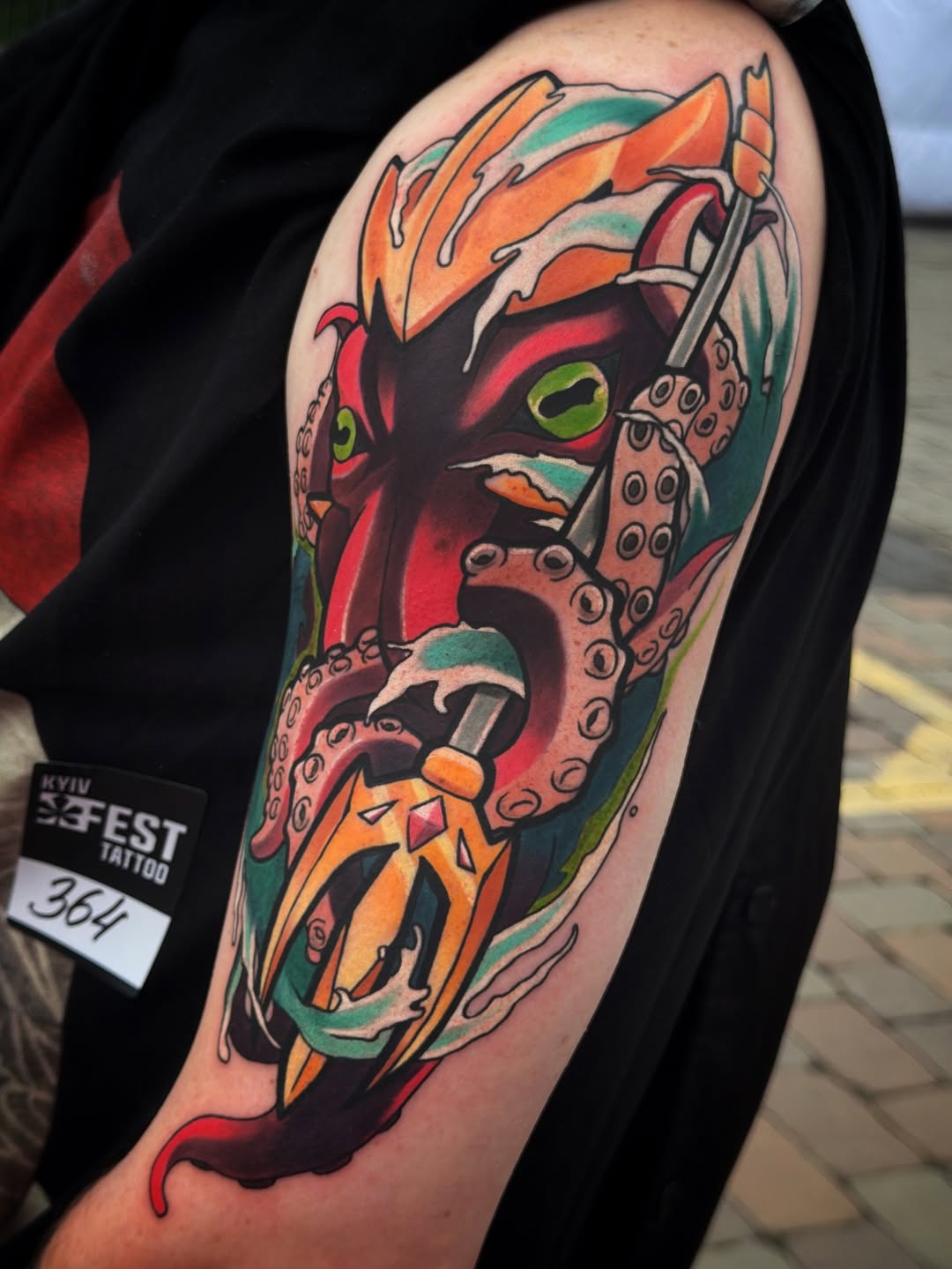 Colorful upper arm tattoo featuring a demon face with green eyes, orange and red coloring, tentacle-like appendages, and ornamental details.