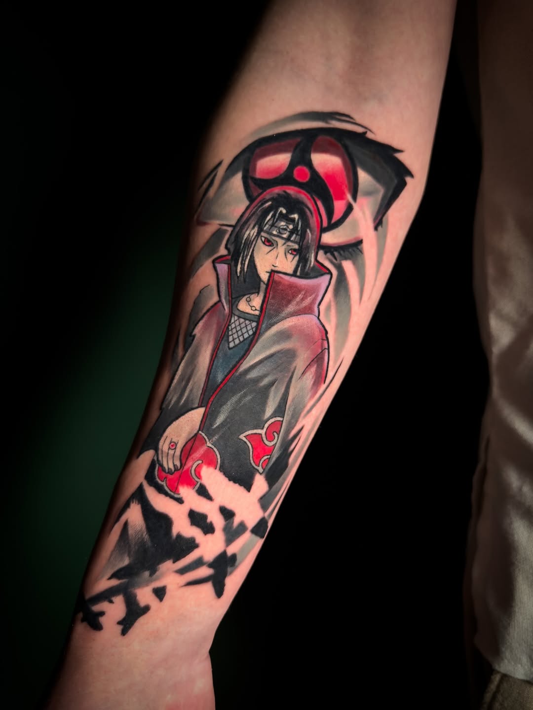 Colored arm tattoo featuring an anime character with long dark hair in red and black clothing, with a large red-eyed symbol above and dark splatter effects below.
