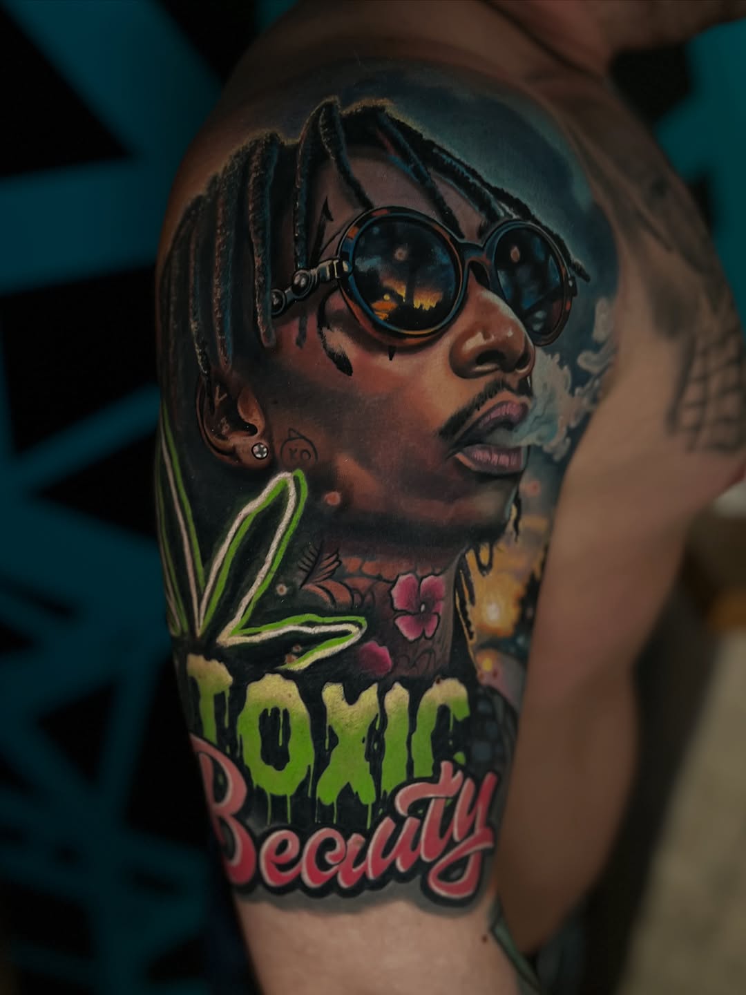 Colorful shoulder tattoo featuring a man with dreadlocks wearing round sunglasses, cannabis leaf, and "Toxic Beauty" text in green and red.