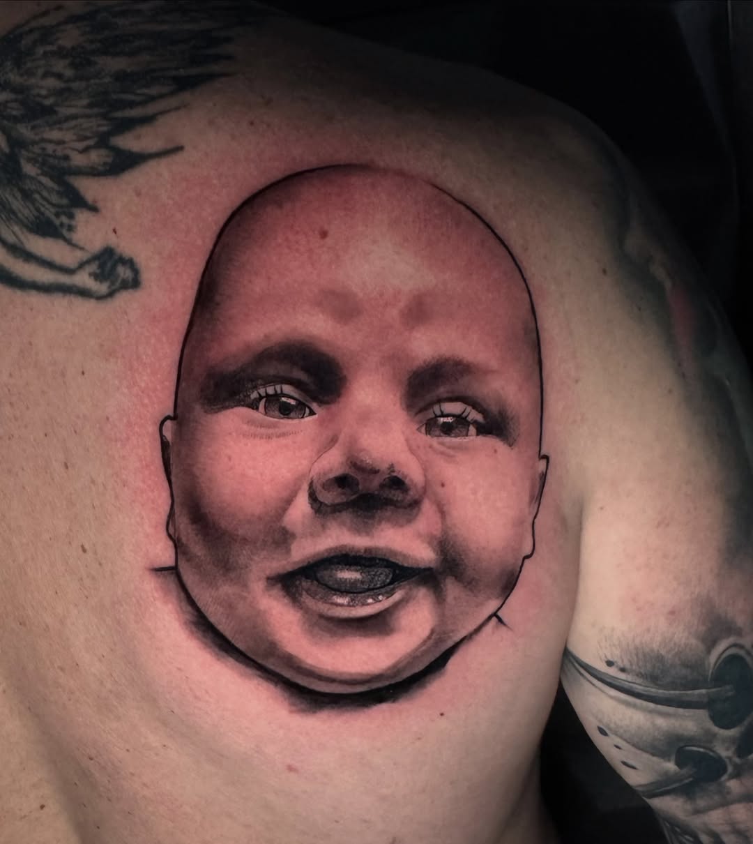 Detailed portrait tattoo of a smiling baby's face with shading on the upper chest and shoulder area.