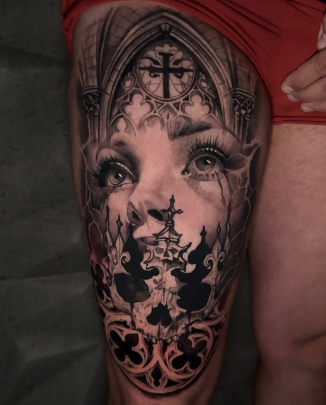 Large thigh tattoo featuring a woman's face merged with a skull, ornate gothic architectural elements, and decorative crosses in black and grey ink.