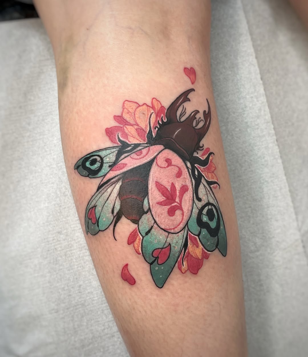 Colorful beetle tattoo on an arm featuring pink and red flowers, teal wings, black antlers, and small hearts, displayed on pale skin.
