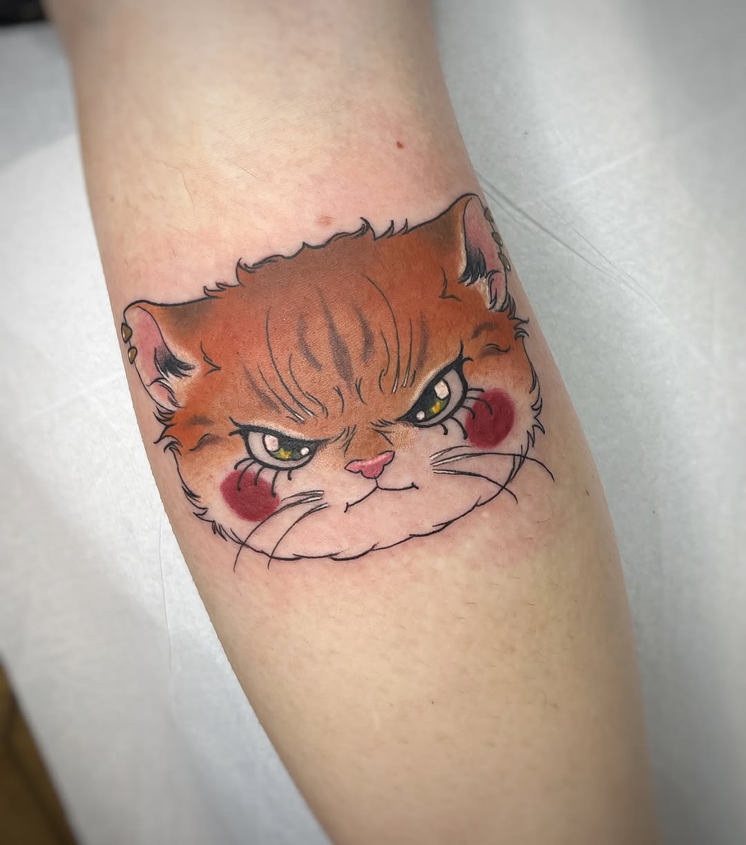 Orange and brown cat face with yellow eyes and rosy cheeks tattooed on a person's forearm with black outlines and pink details.