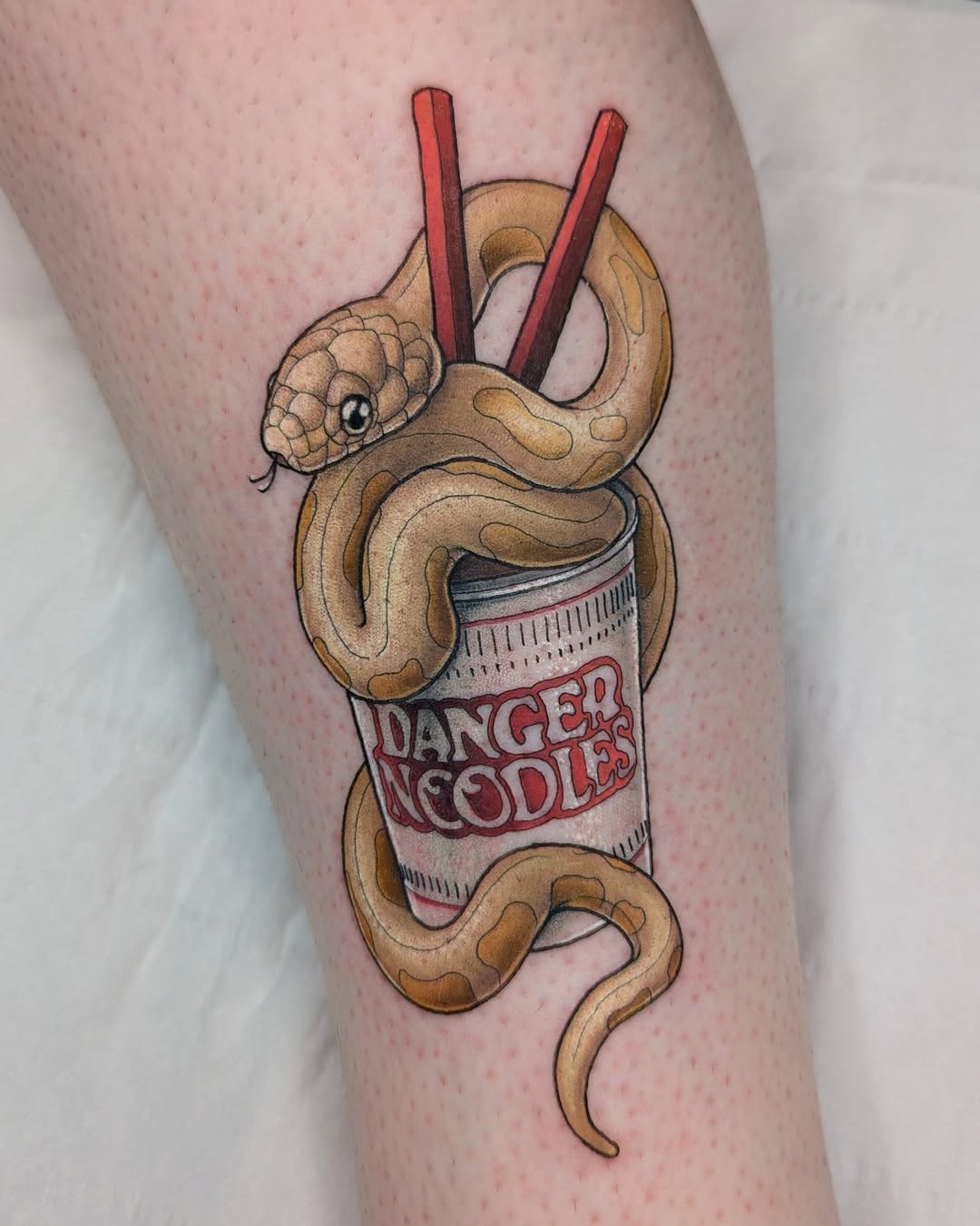 Tan snake wrapped around a noodle container labeled "Danger Noodles" with red chopsticks, tattooed on a leg in brown, red, and white colors.