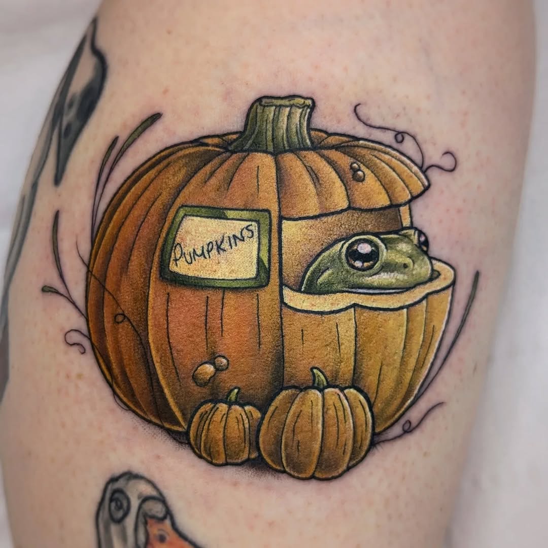 Colorful pumpkin tattoo on upper arm featuring a frog peeking from the gourd, smaller pumpkins below, and a "Pumpkins" label, with green vines and orange-yellow shading.