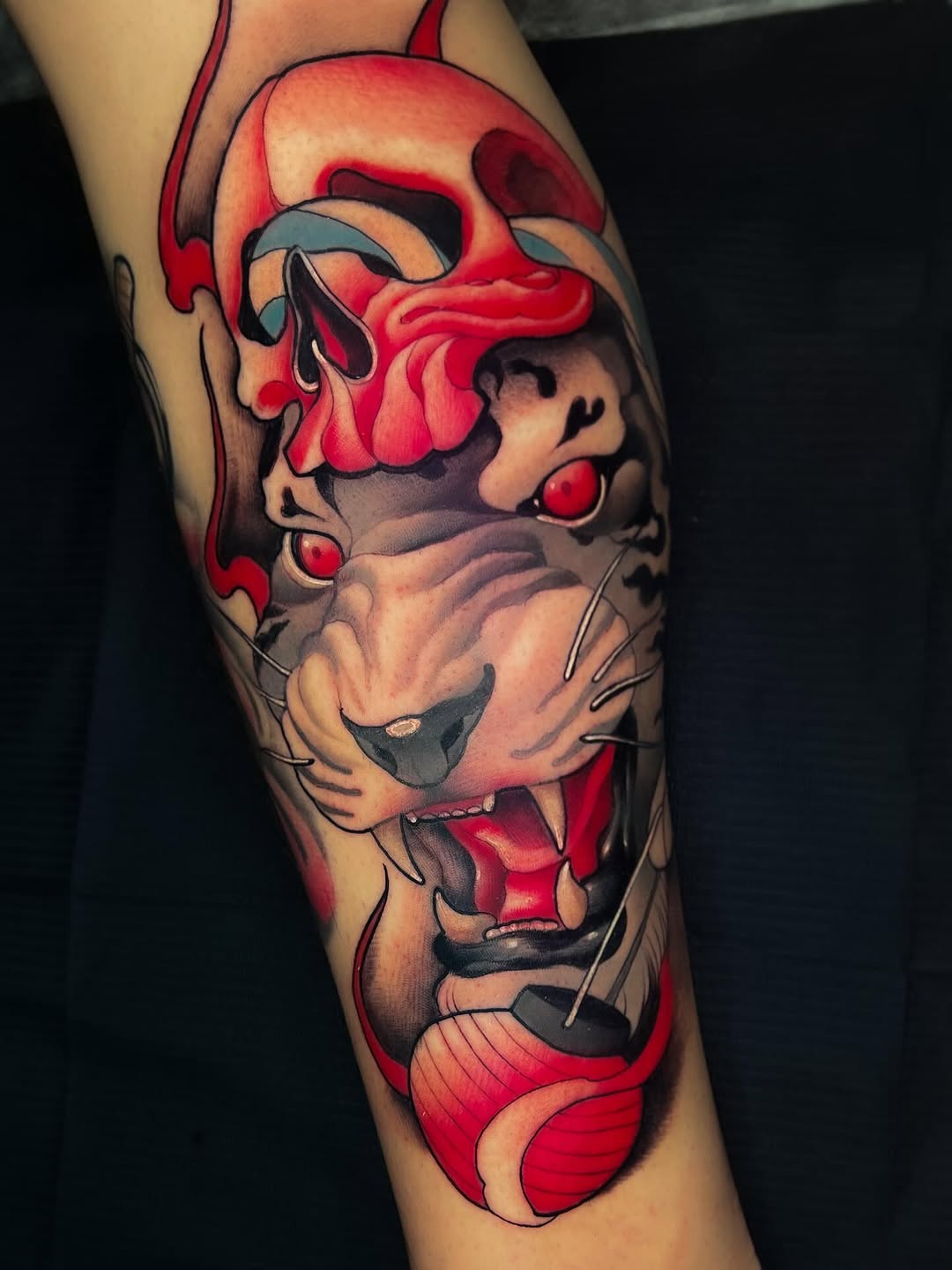 Colorful tattoo on a leg featuring a stylized face with red and gray tones, intricate line work, and expressive features surrounded by flowing red shapes.