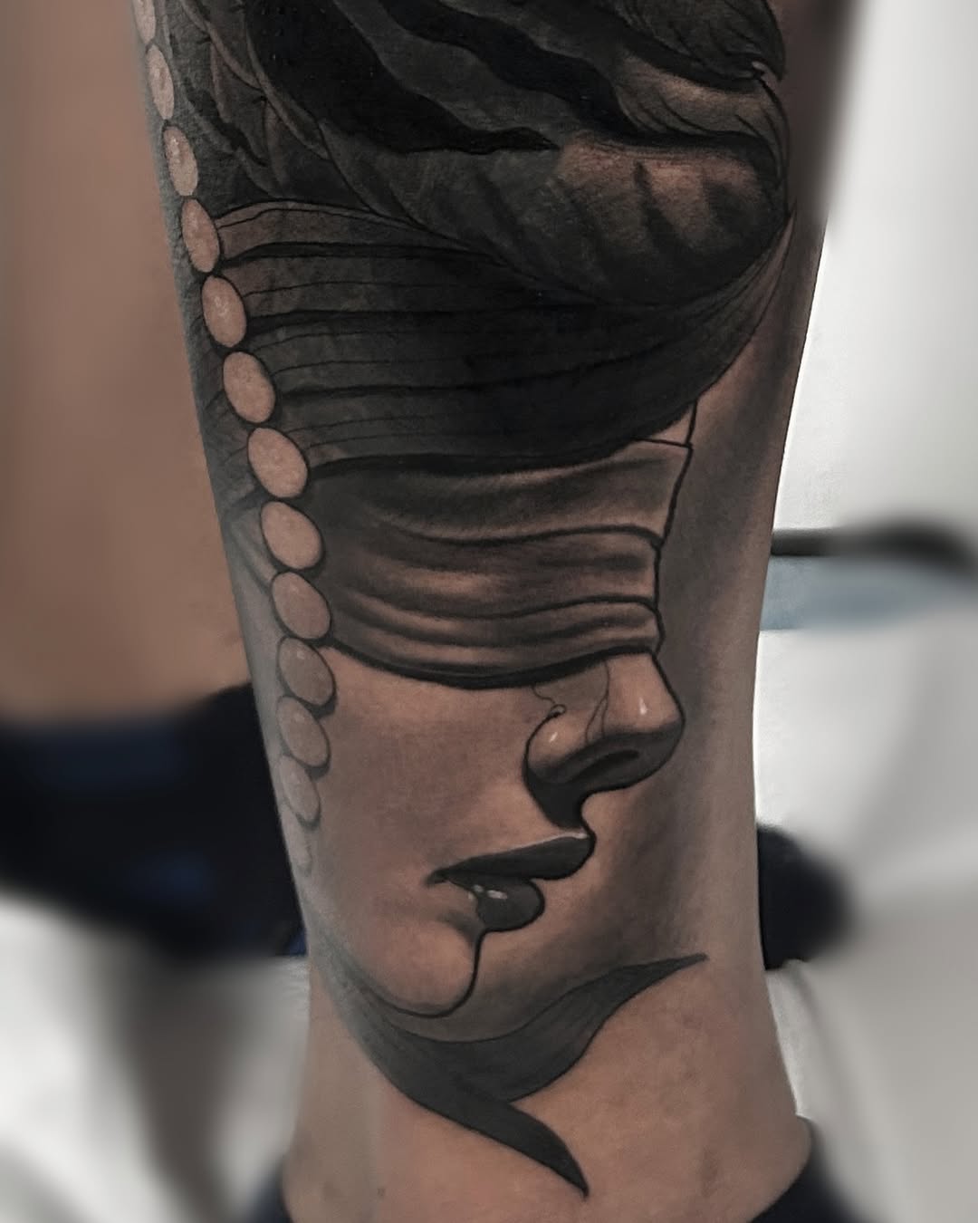 Close-up side profile portrait tattoo on forearm showing a face with closed eyes, lips, and decorative circular elements along the upper shoulder area in black and grey ink.