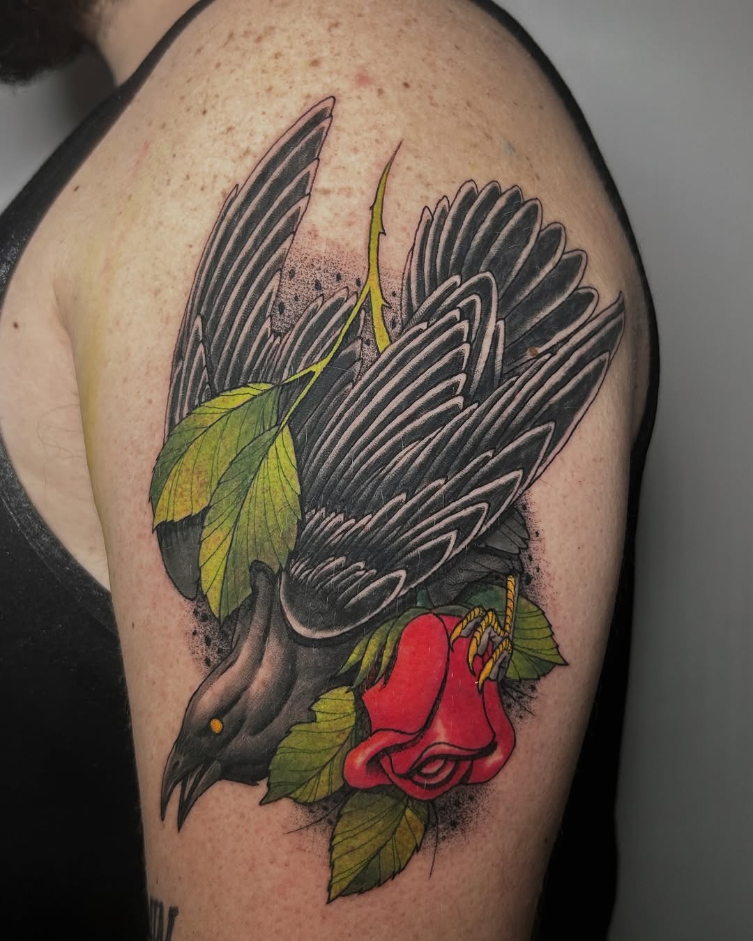 A dark bird with spread wings holding a red rose with green leaves in its beak, tattooed on the upper arm.