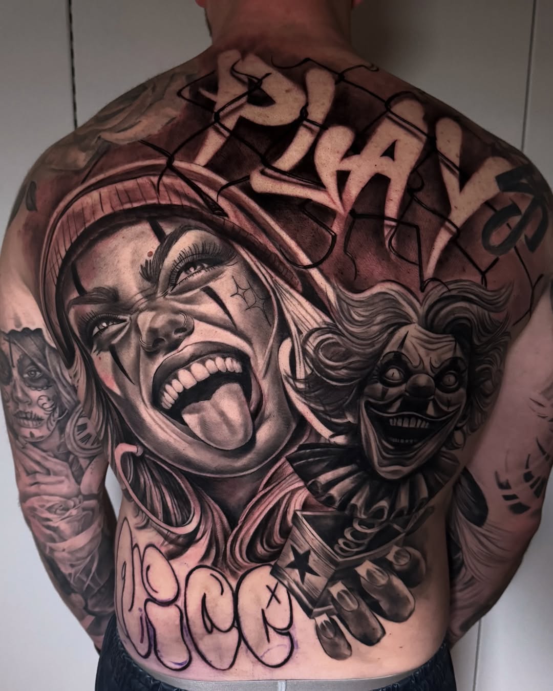 Large back tattoo featuring two expressive faces with detailed shading, ornamental elements, and text script in black and gray ink.