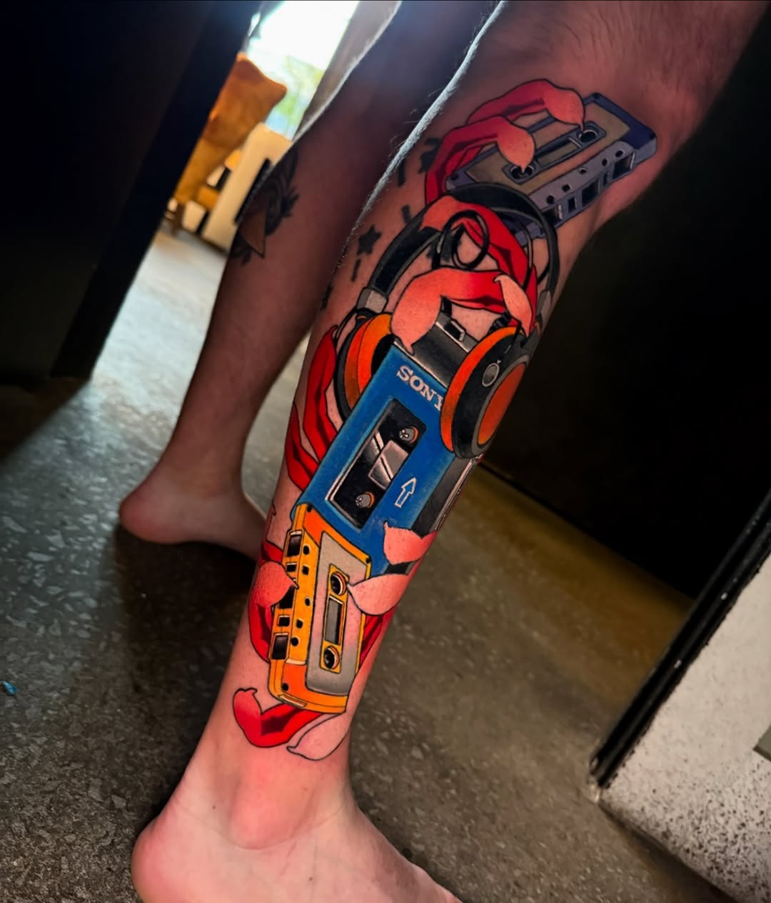 Colorful stacked cassette tape tattoo on a leg featuring red, blue, and yellow Sony and other branded tapes with decorative red ribbon elements.