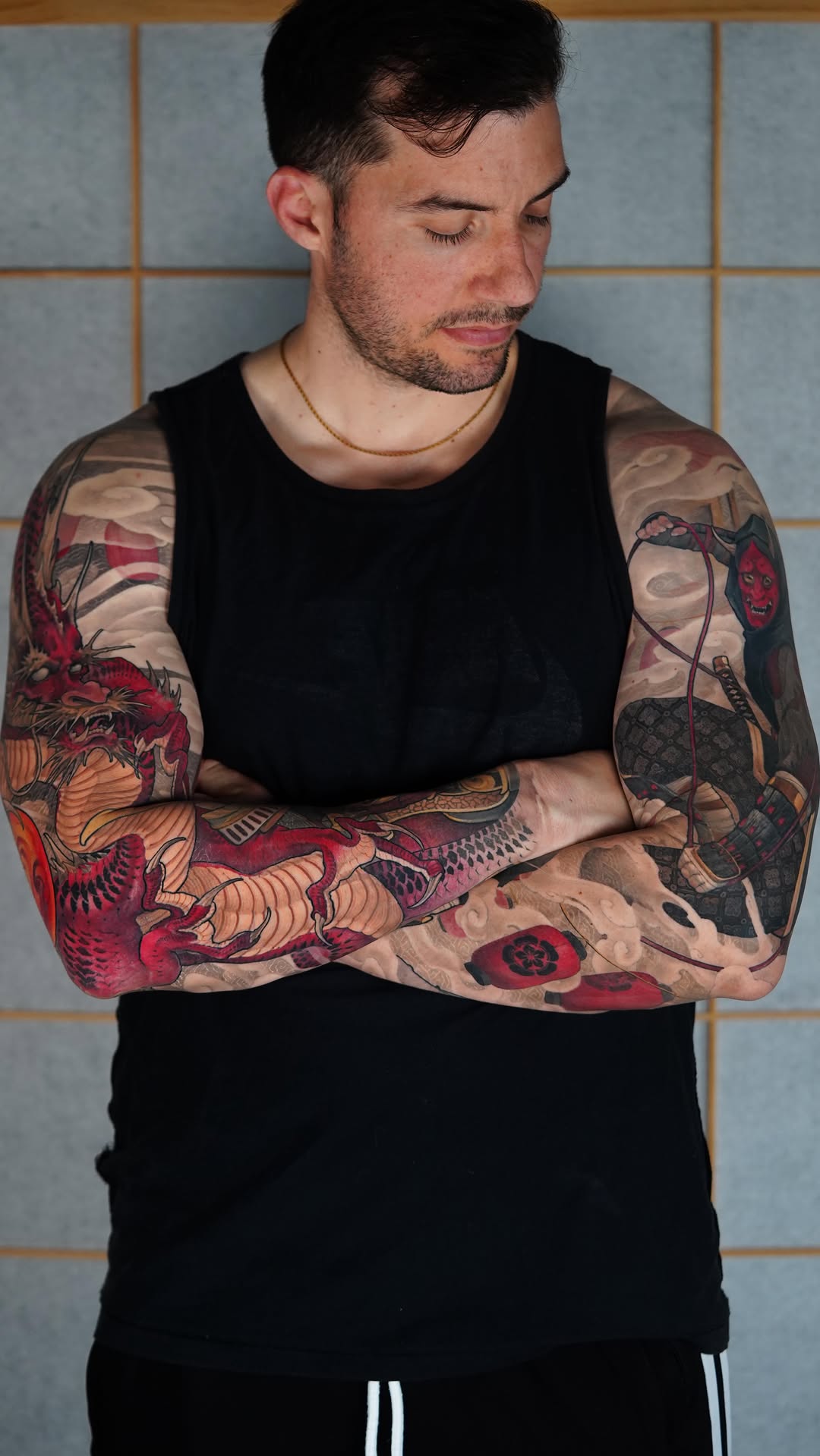 Man displaying full-sleeve arm tattoos featuring red and orange creatures, demons, and mythological figures with detailed linework against a tiled background.