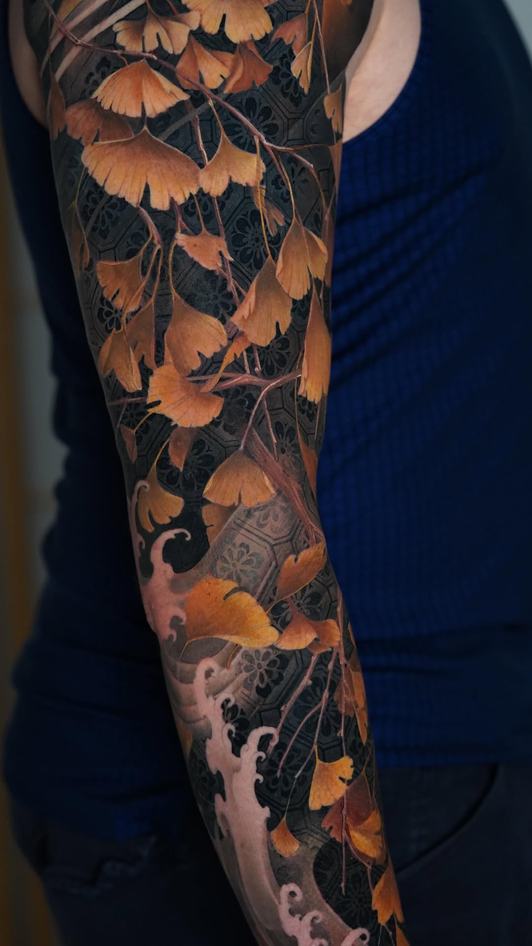 Full-length arm sleeve tattoo featuring golden ginkgo leaves cascading down dark ornamental patterned background in orange, black, and brown tones.