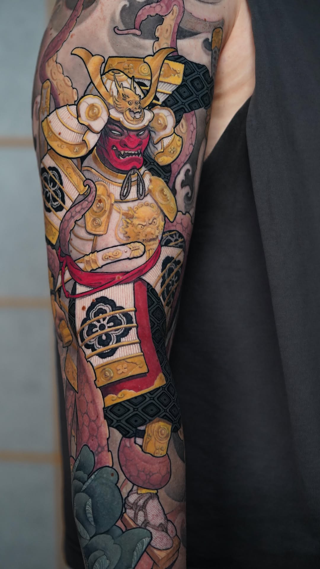Colorful arm tattoo featuring a red-faced demon warrior with gold armor, horns, and ornate decorative elements surrounded by mythological symbols and imagery.