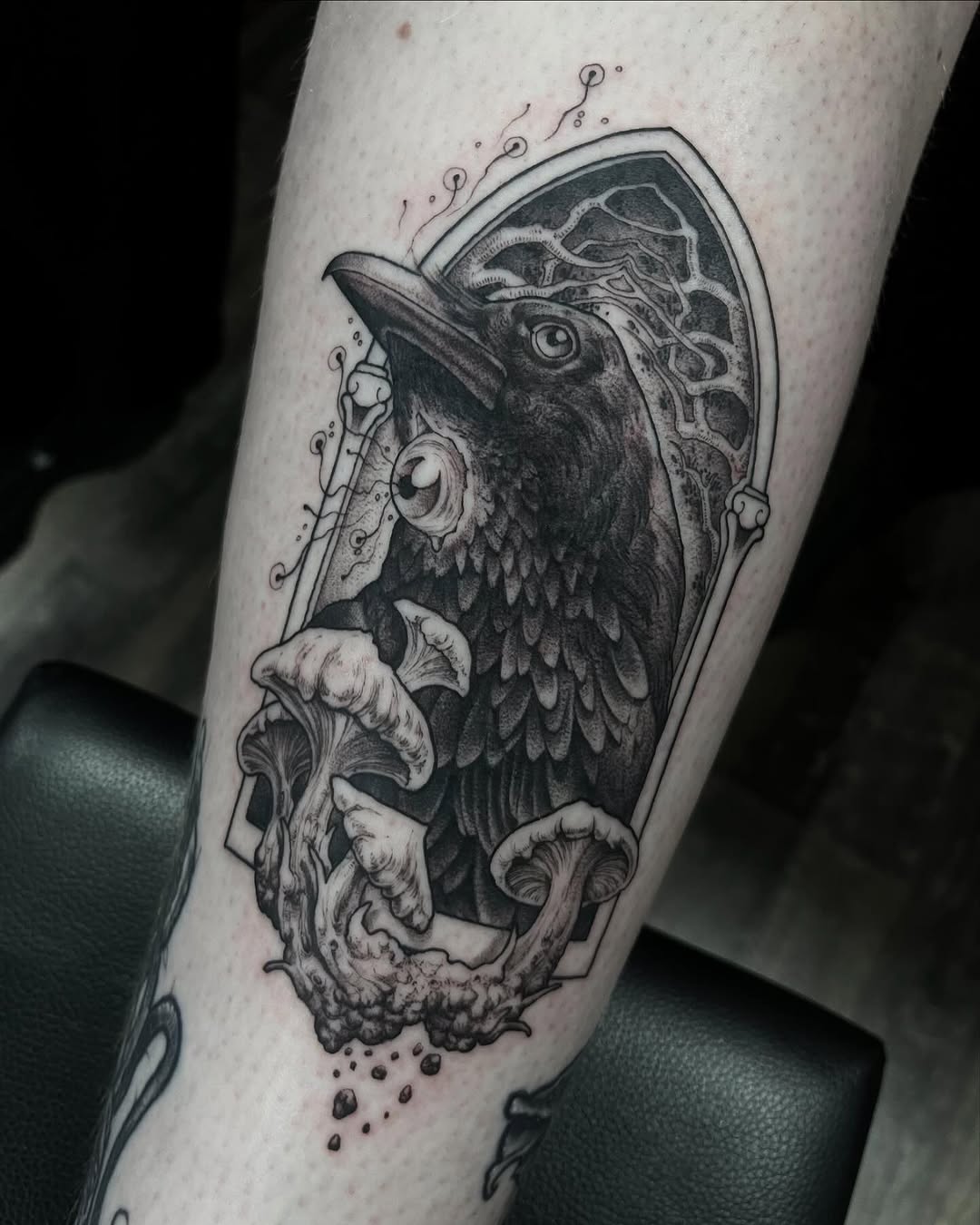 Raven's head in ornate frame with mushrooms and decorative flourishes tattooed on upper leg in black ink.